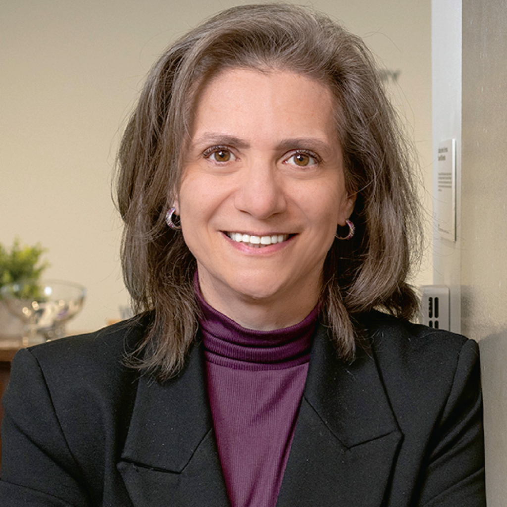 A person with shoulder-length brown hair, wearing a black blazer and purple turtleneck, smiles while standing indoors near a light-colored wall.