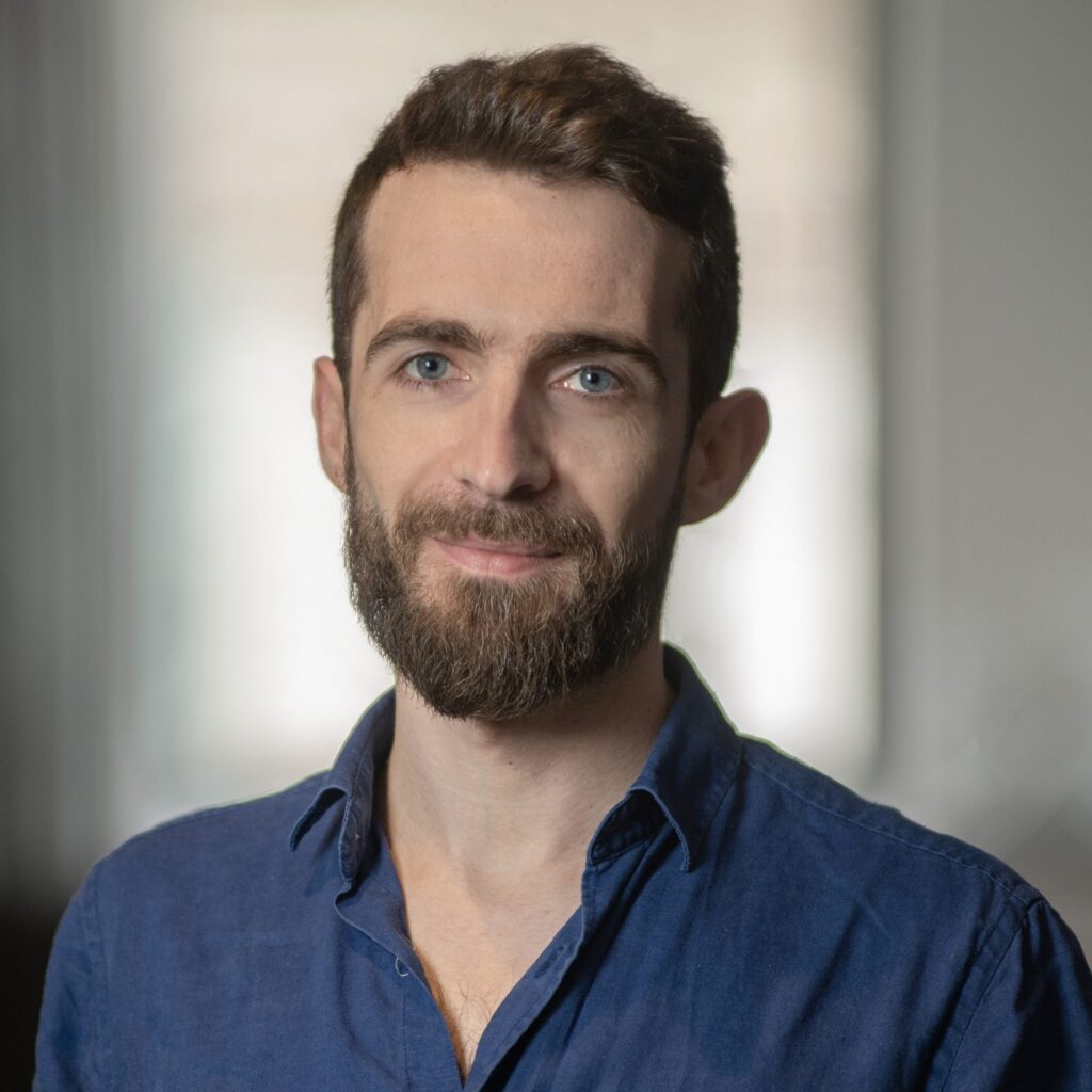 A man with short brown hair and a beard, wearing a blue shirt, stands indoors with a neutral expression. The background is softly blurred.