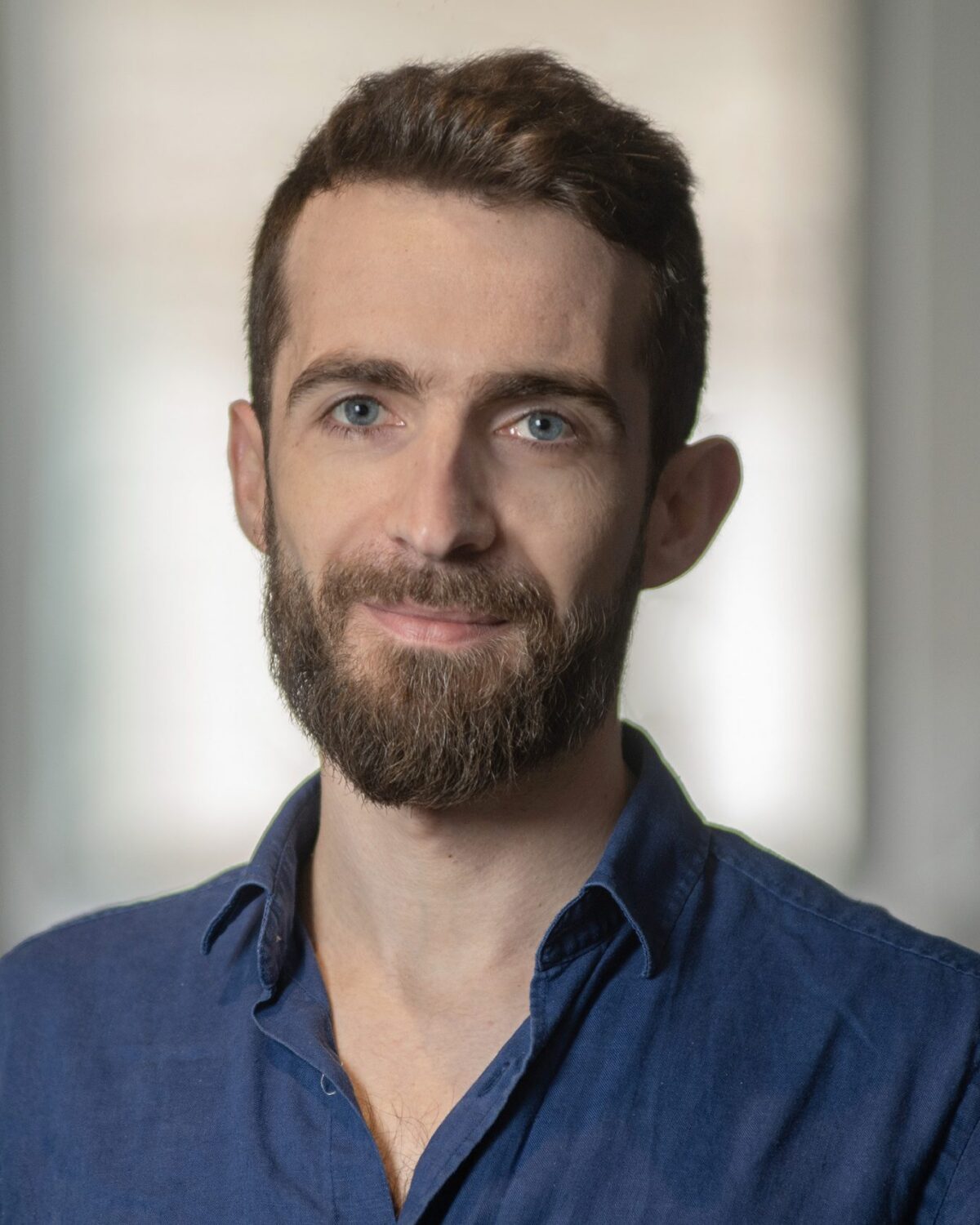 A man with short brown hair and a beard, wearing a blue shirt, stands indoors with a neutral expression. The background is softly blurred.