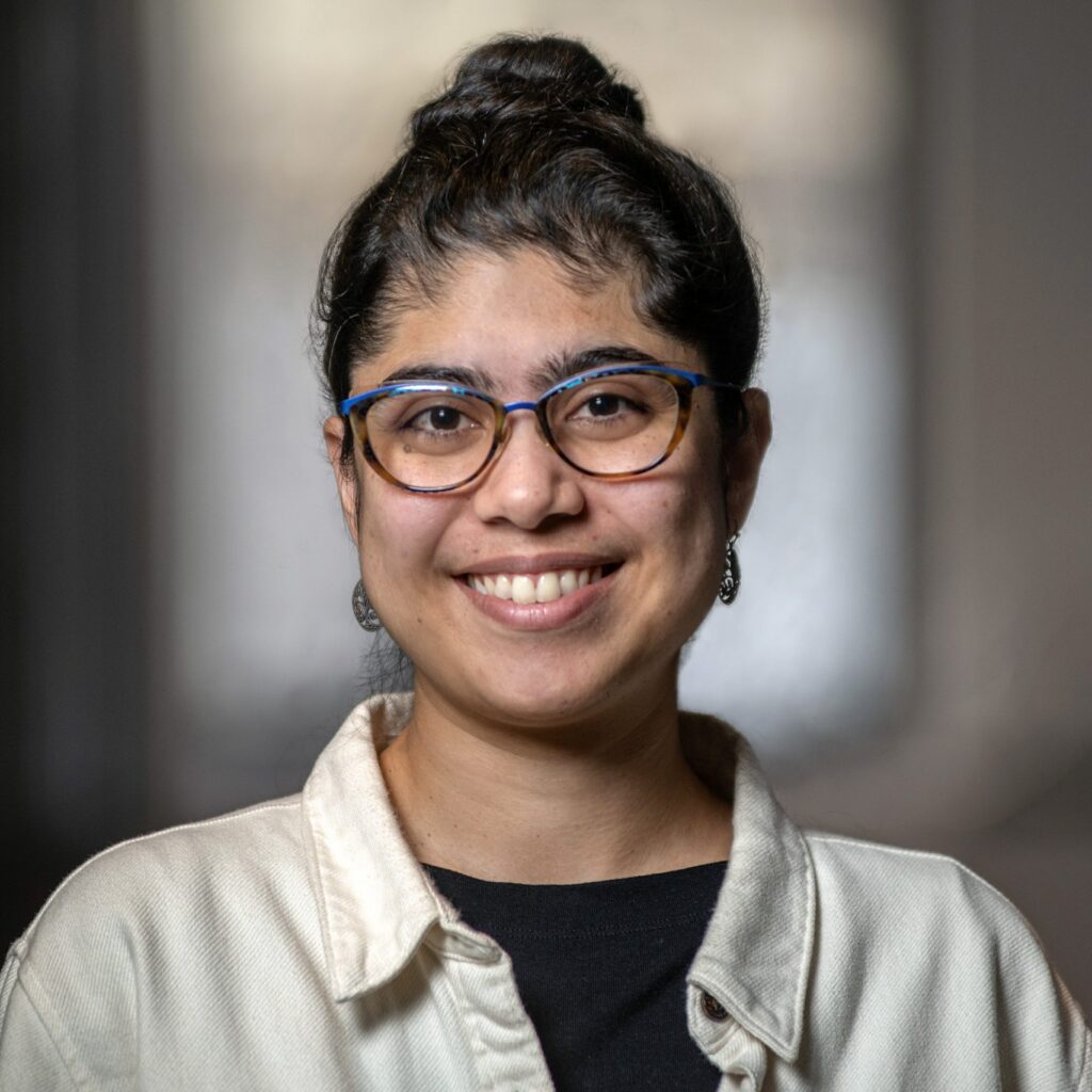 A person with long dark hair in a bun, wearing blue glasses, earrings, a cream jacket, and a black shirt, smiles at the camera with a blurred indoor background.