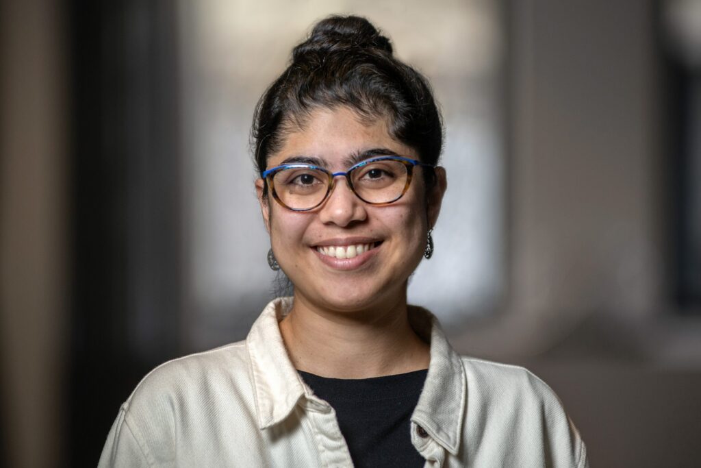A person with long dark hair in a bun, wearing blue glasses, earrings, a cream jacket, and a black shirt, smiles at the camera with a blurred indoor background.