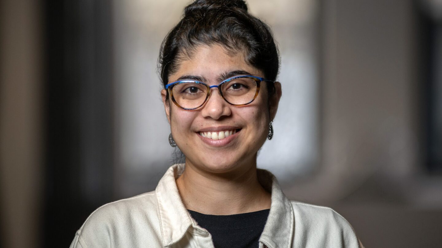 A person with long dark hair in a bun, wearing blue glasses, earrings, a cream jacket, and a black shirt, smiles at the camera with a blurred indoor background.