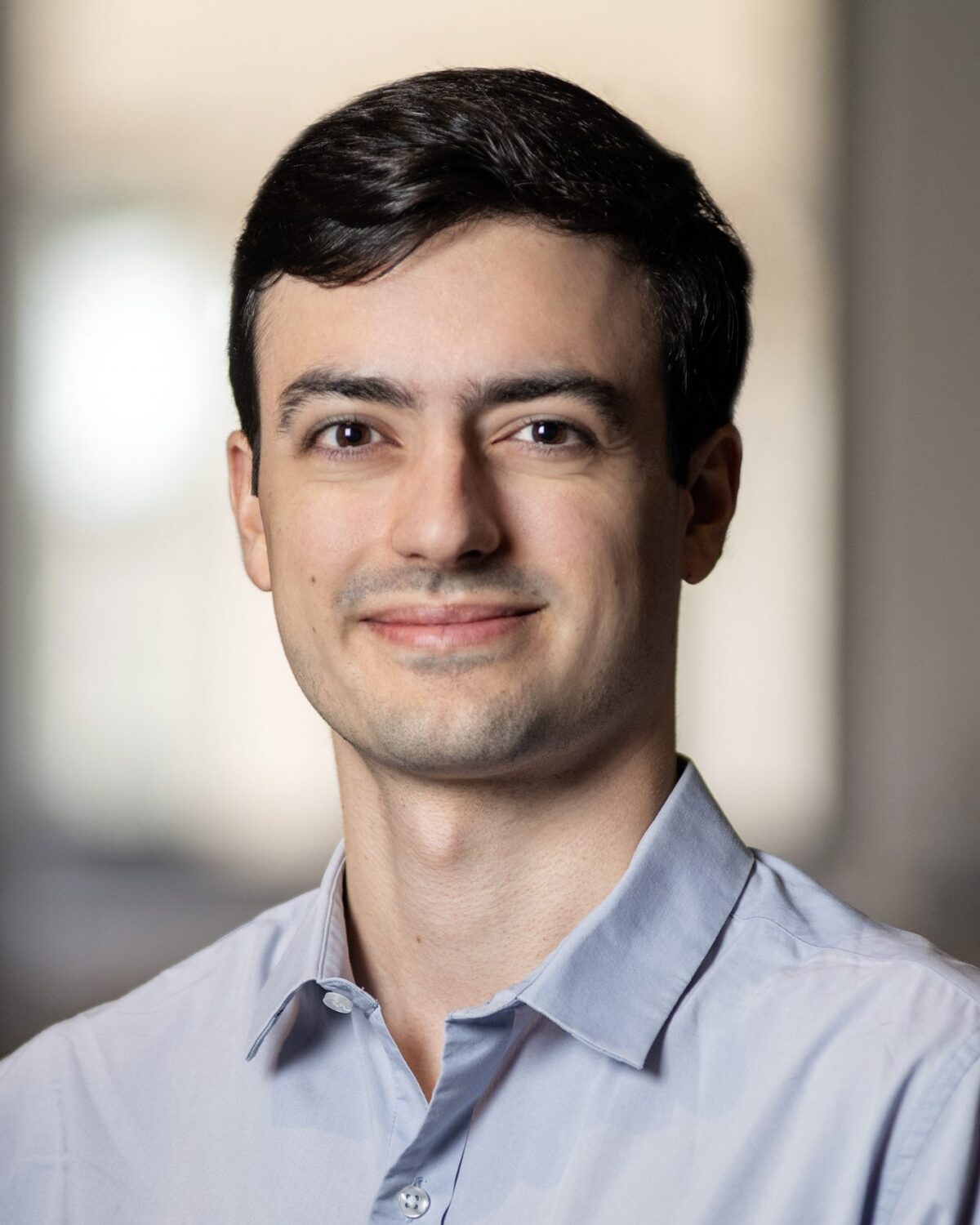 A young man with short dark hair, wearing a light blue button-up shirt, smiles softly while looking at the camera. The background is softly blurred with windows visible.