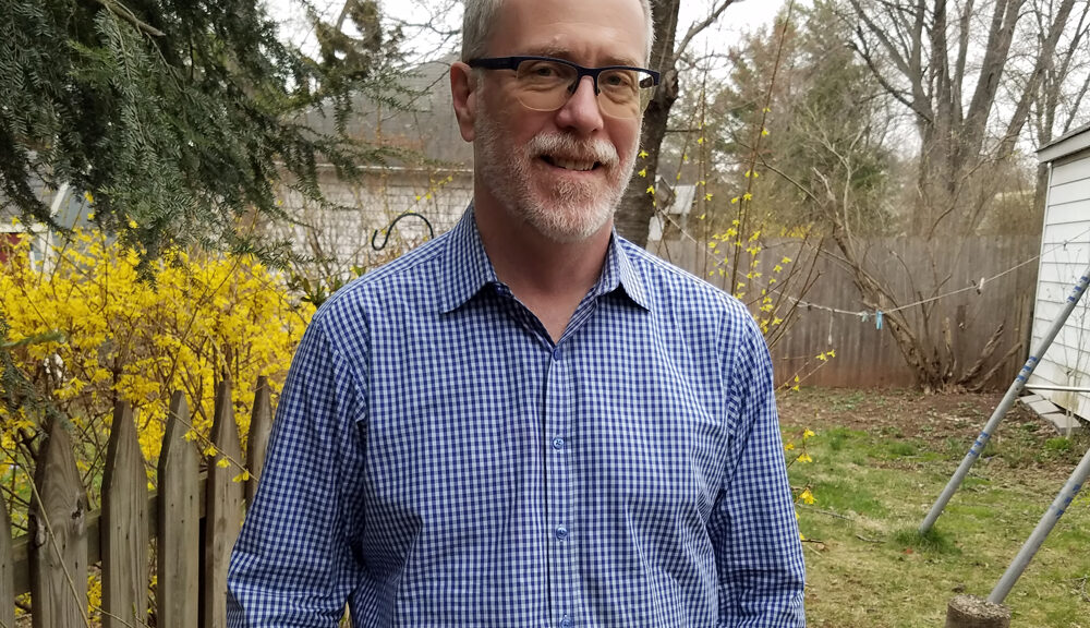 A man with glasses and a beard stands outside by a wooden fence, wearing a blue checkered shirt. Yellow flowers and trees are in the background on a cloudy day.