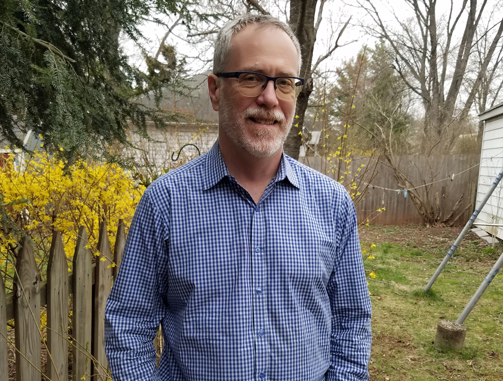 A man with glasses and a beard stands outside by a wooden fence, wearing a blue checkered shirt. Yellow flowers and trees are in the background on a cloudy day.