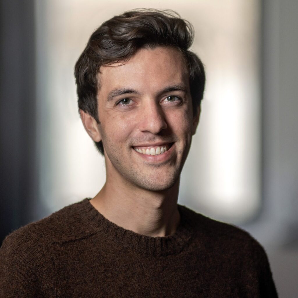 A man with short brown hair, wearing a brown sweater, smiles at the camera in a softly lit indoor setting with a blurred background.