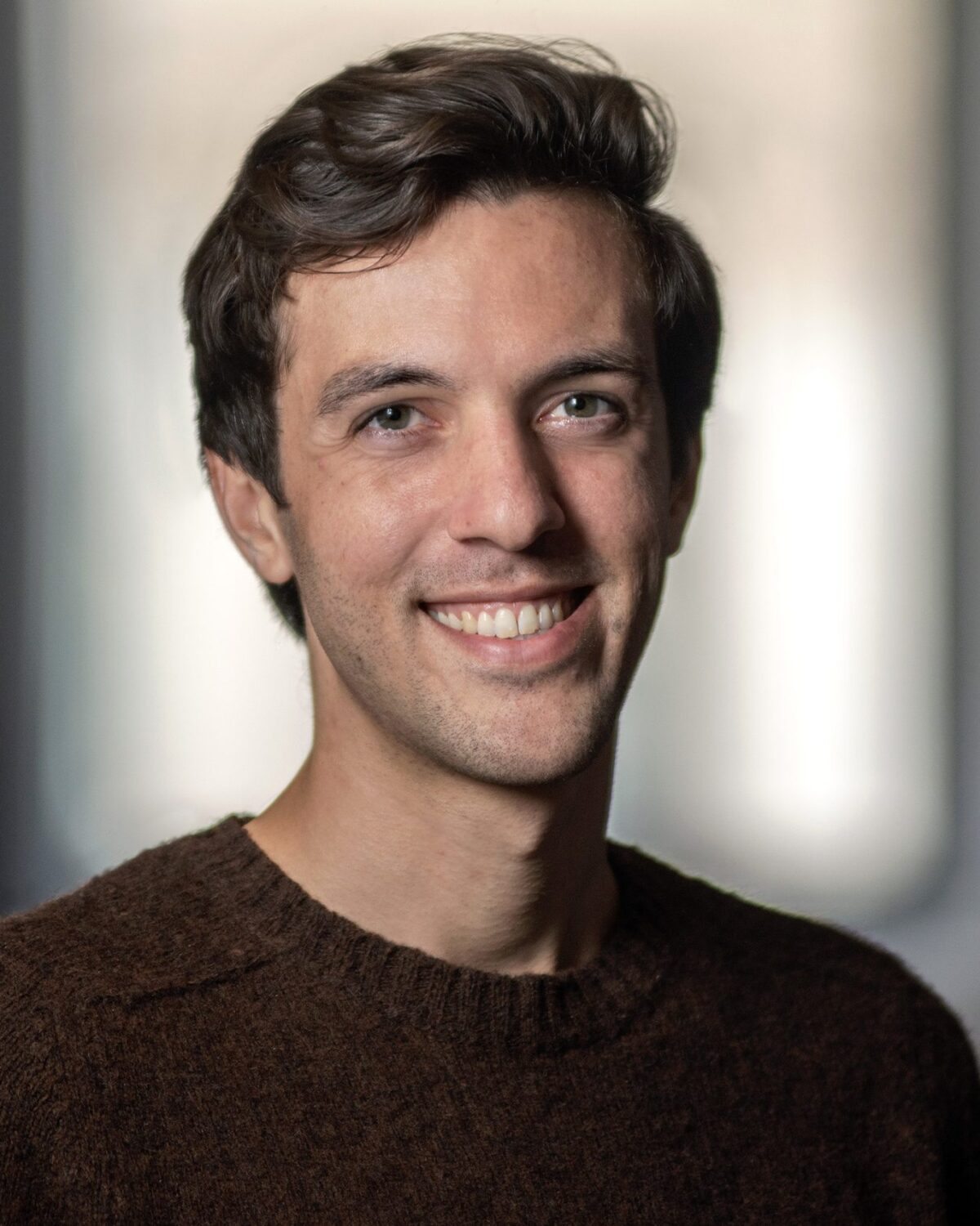 A man with short brown hair, wearing a brown sweater, smiles at the camera in a softly lit indoor setting with a blurred background.
