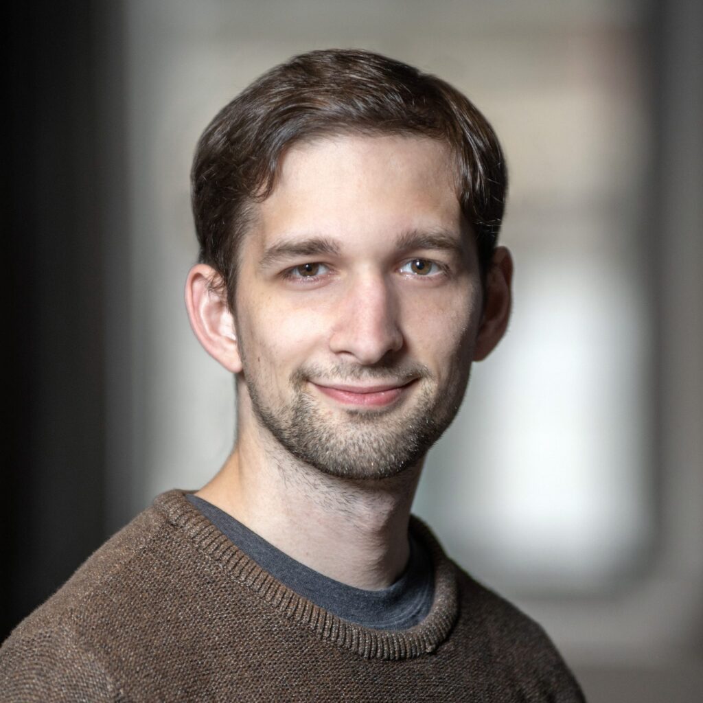 A young man with short brown hair and a beard, wearing a brown sweater, smiles gently at the camera. The background is softly blurred, highlighting his face.