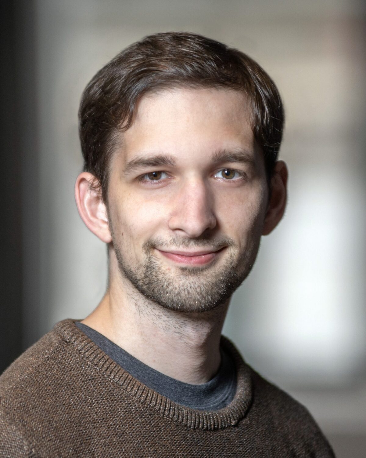 A young man with short brown hair and a beard, wearing a brown sweater, smiles gently at the camera. The background is softly blurred, highlighting his face.