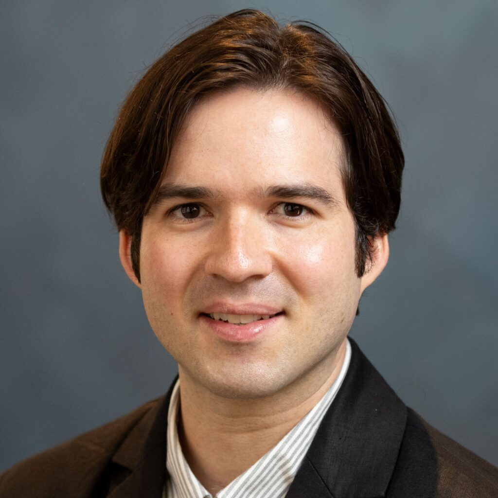 A person with medium-length brown hair, fair skin, and dark eyes is wearing a dark blazer over a light shirt, smiling slightly in front of a blurred blue-gray background.