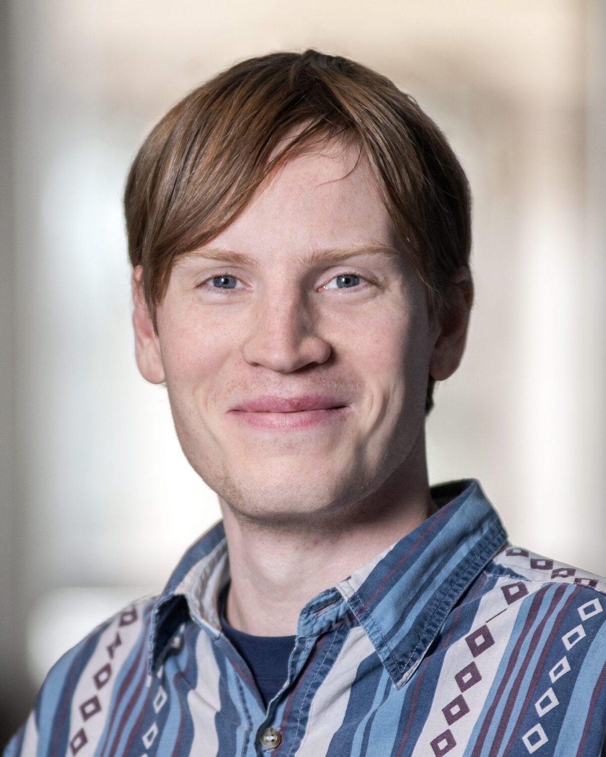 A person with light skin and straight, light brown hair smiles softly while wearing a patterned button-up shirt. The background is softly blurred, focusing attention on the individual.