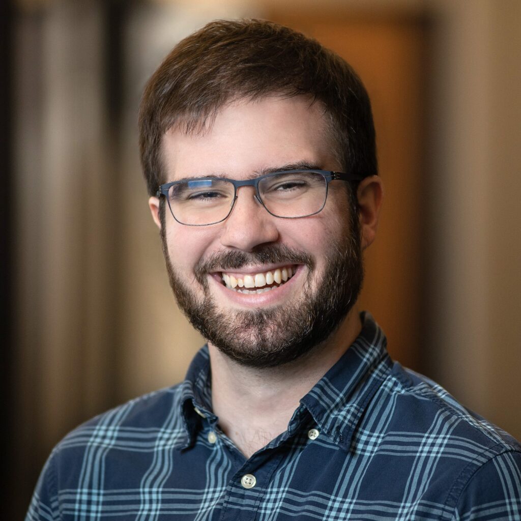 A man with short brown hair, glasses, and a beard smiles widely while wearing a blue plaid shirt, standing in a softly blurred indoor setting.