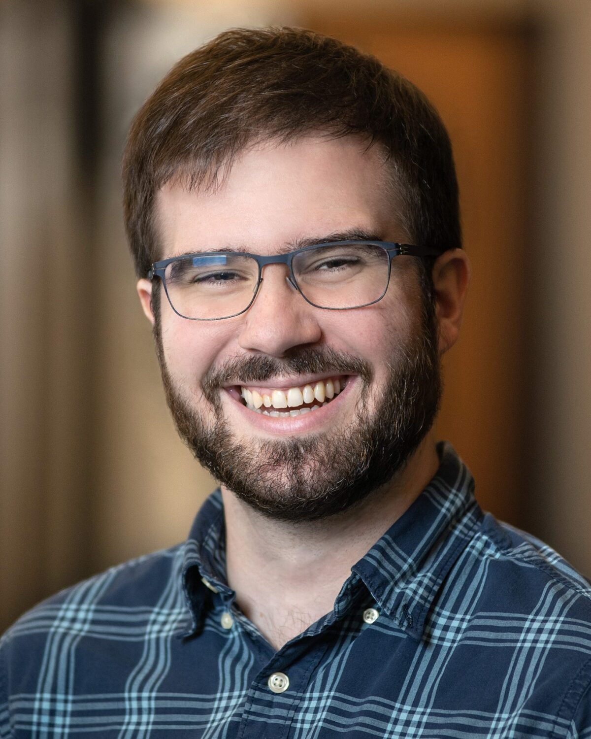 A man with short brown hair, glasses, and a beard smiles widely while wearing a blue plaid shirt, standing in a softly blurred indoor setting.