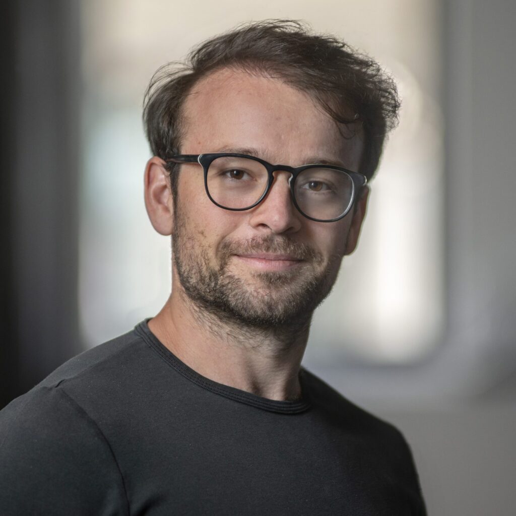 A man with short brown hair, glasses, and a beard is wearing a dark shirt and looking at the camera, smiling slightly. The background is blurred.