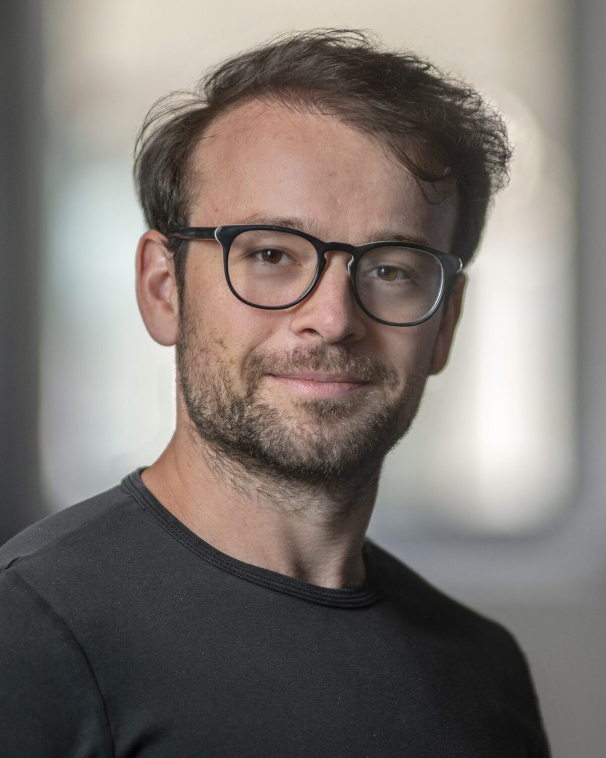 A man with short brown hair, glasses, and a beard is wearing a dark shirt and looking at the camera, smiling slightly. The background is blurred.