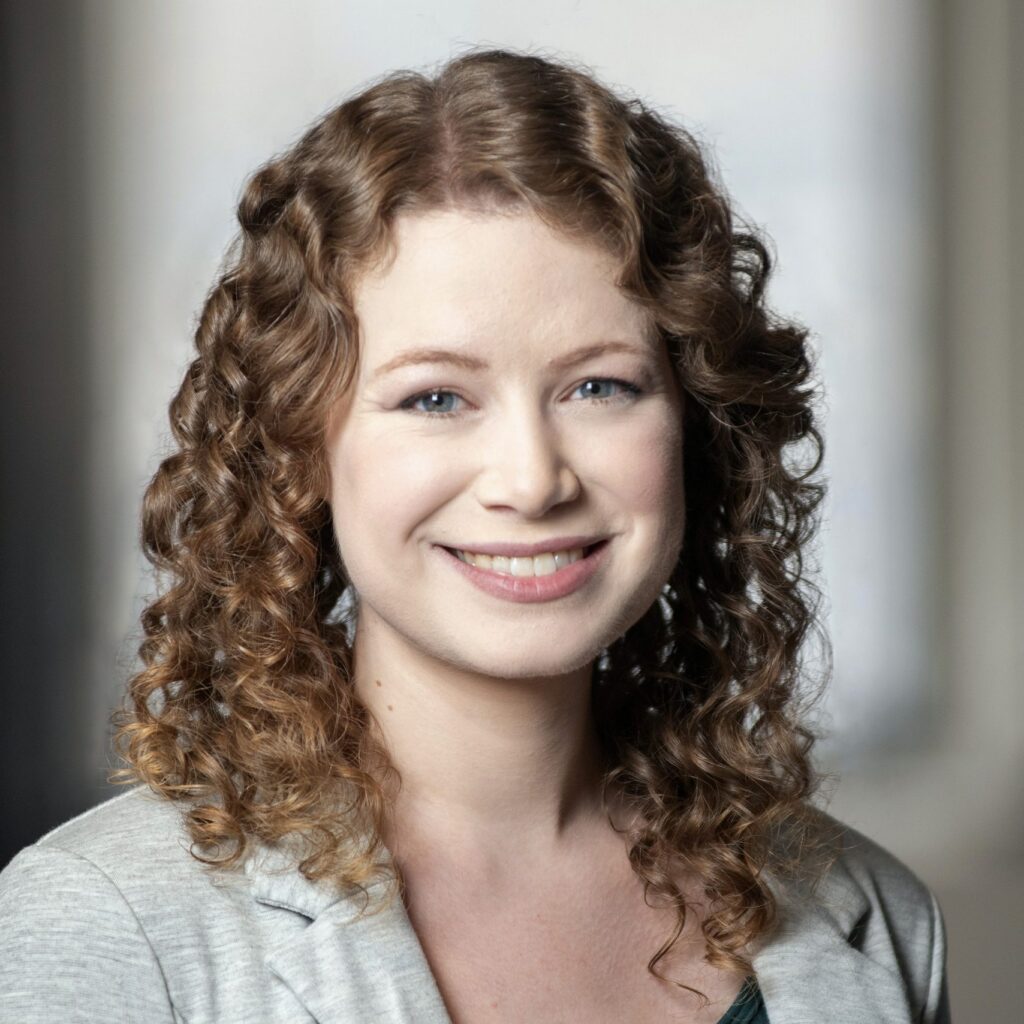A woman with light skin and curly, light brown hair smiles at the camera. She is wearing a light gray blazer over a dark top. The background is softly blurred.