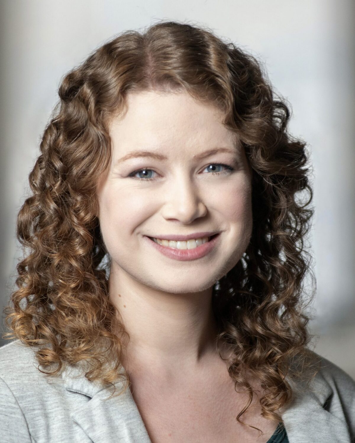A woman with light skin and curly, light brown hair smiles at the camera. She is wearing a light gray blazer over a dark top. The background is softly blurred.