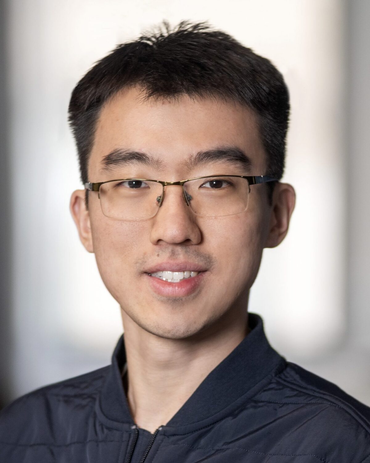 A young man wearing glasses and a dark jacket smiles at the camera, with a softly blurred indoor background.