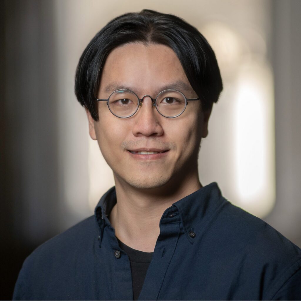 A person with medium-length black hair, round glasses, and a dark blue shirt is smiling slightly while looking at the camera. The background is softly blurred.