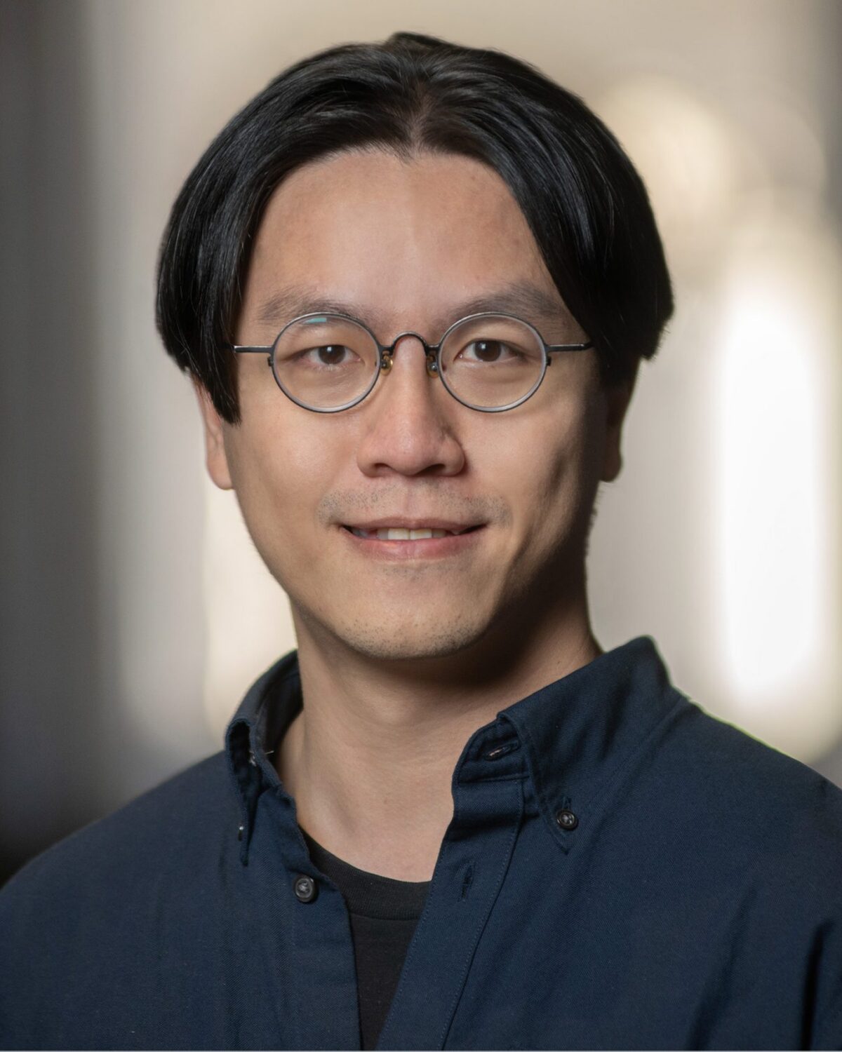 A person with medium-length black hair, round glasses, and a dark blue shirt is smiling slightly while looking at the camera. The background is softly blurred.