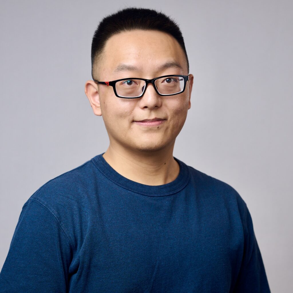 A person wearing glasses and a blue t-shirt stands in front of a plain light gray background, smiling slightly with short black hair.