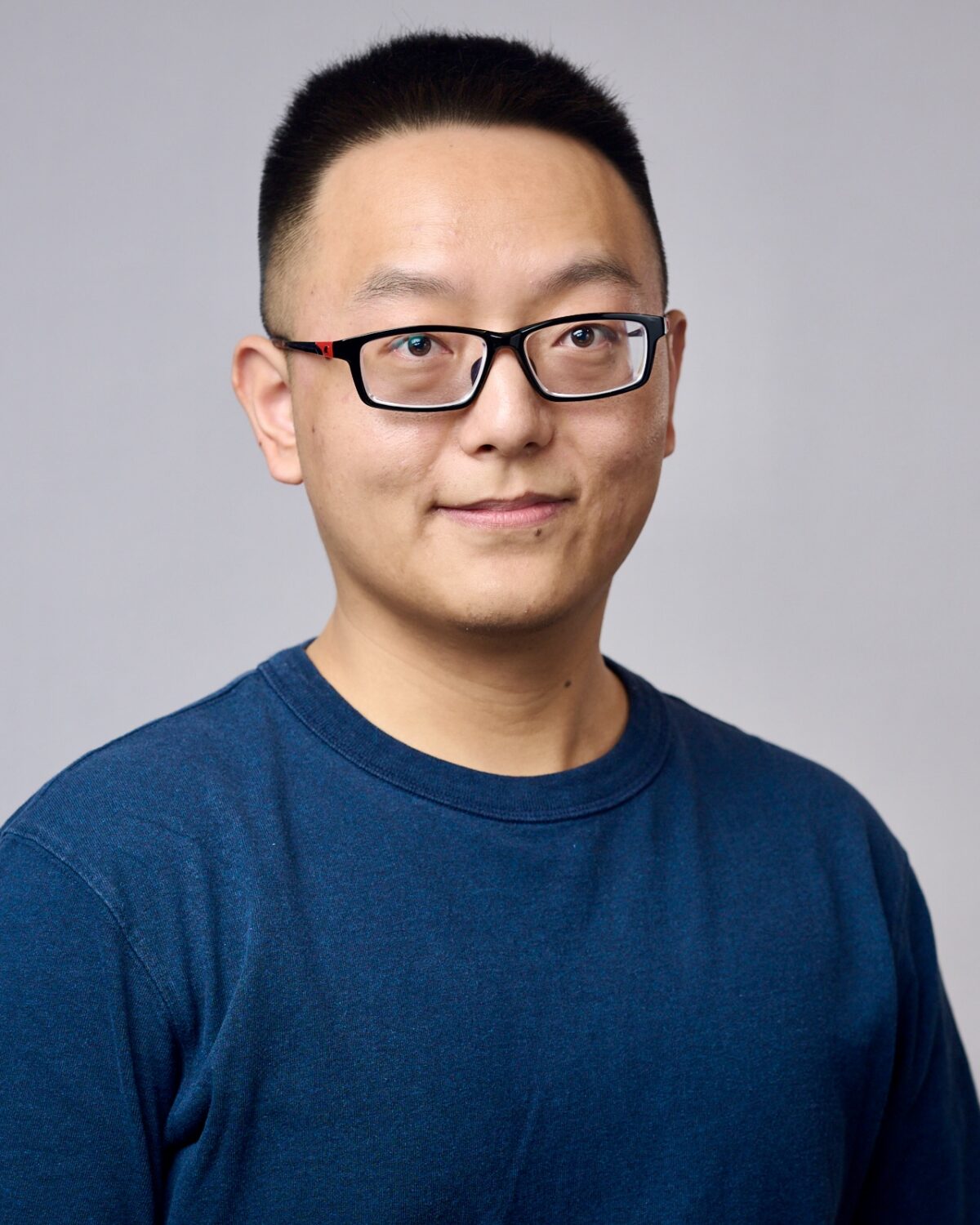 A person wearing glasses and a blue t-shirt stands in front of a plain light gray background, smiling slightly with short black hair.