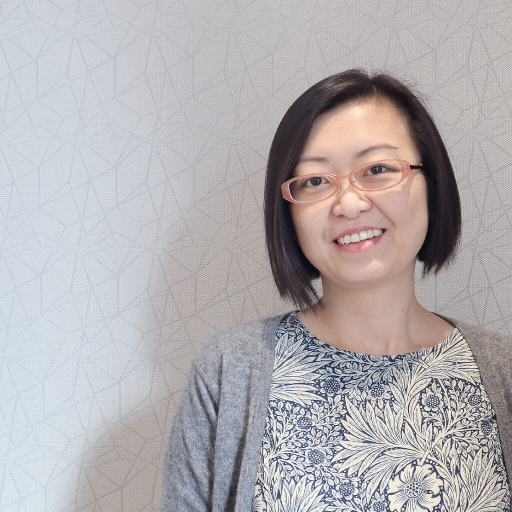 A woman with short black hair and glasses smiles at the camera, standing against a light-colored, patterned wall. She wears a floral-patterned top and a gray cardigan.