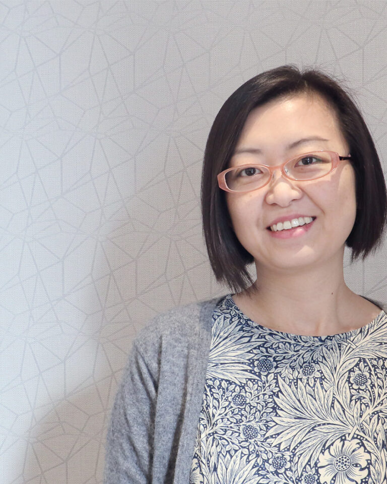 A woman with short black hair and glasses smiles at the camera, standing against a light-colored, patterned wall. She wears a floral-patterned top and a gray cardigan.