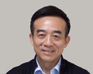 A middle-aged man with short black hair, wearing a collared shirt and dark jacket, smiles at the camera against a plain light background.
