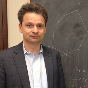 A man in a suit stands in front of a chalkboard with geometric drawings and mathematical notations.