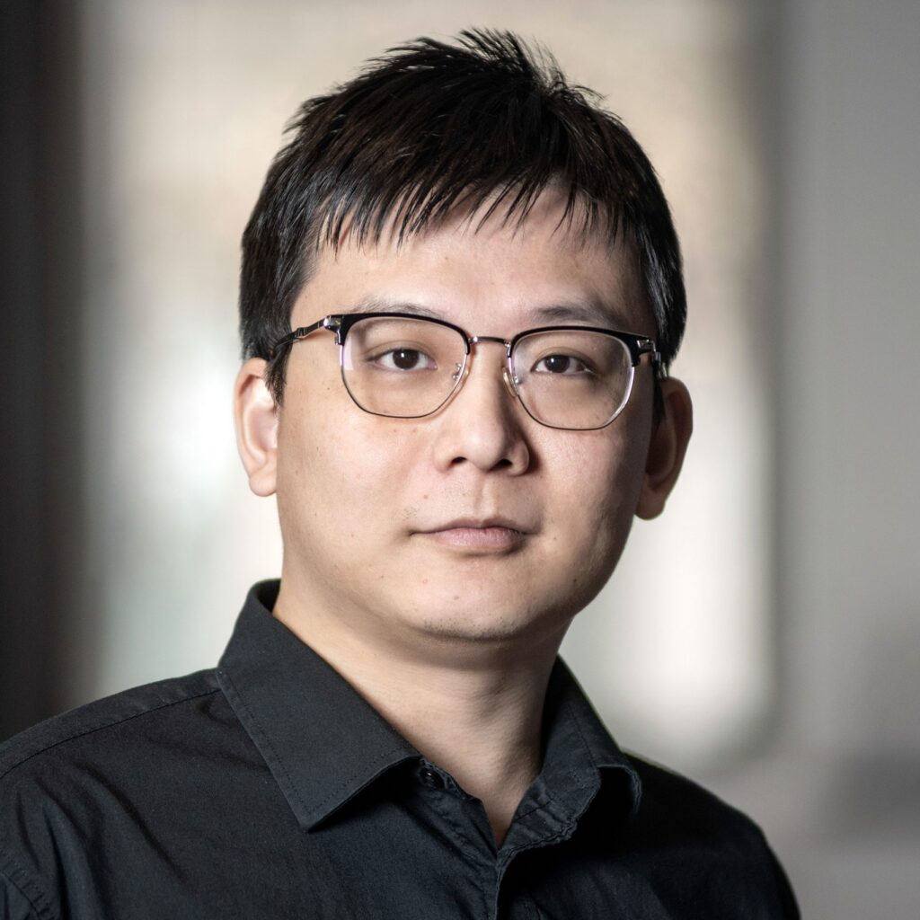 A person with short dark hair and glasses wearing a black collared shirt stands indoors in front of a blurred background, looking at the camera with a neutral expression.