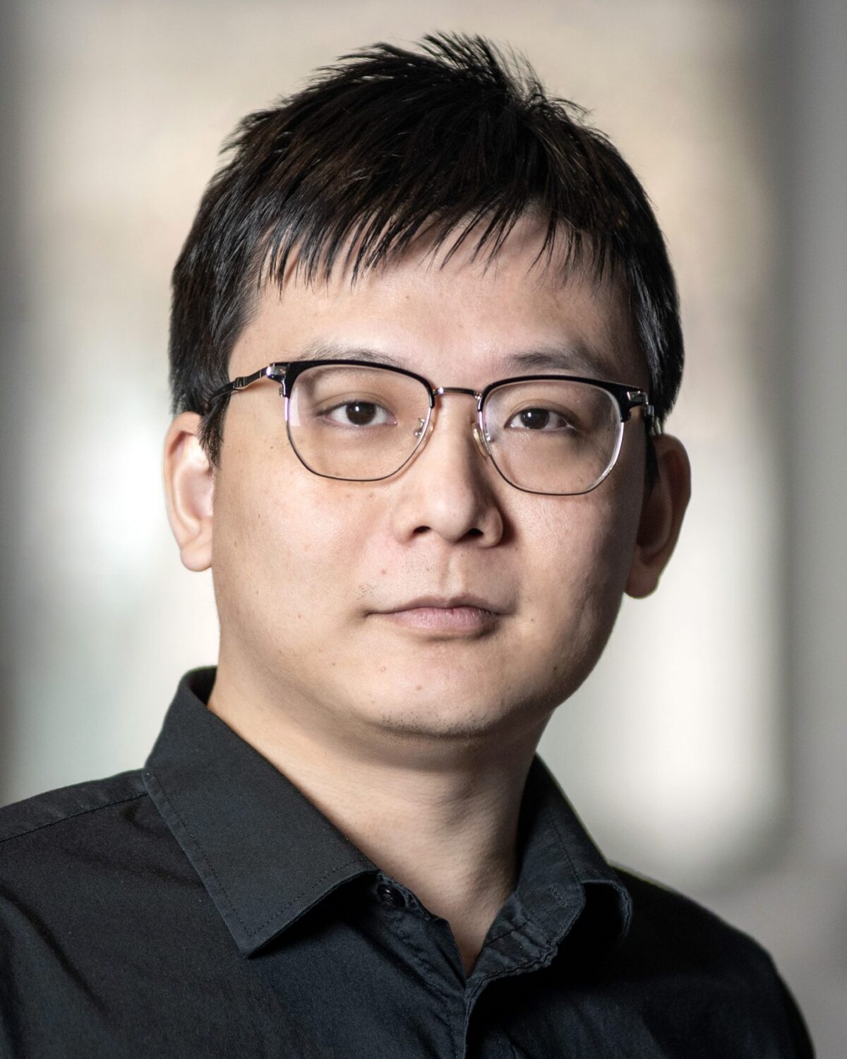 A person with short dark hair and glasses wearing a black collared shirt stands indoors in front of a blurred background, looking at the camera with a neutral expression.
