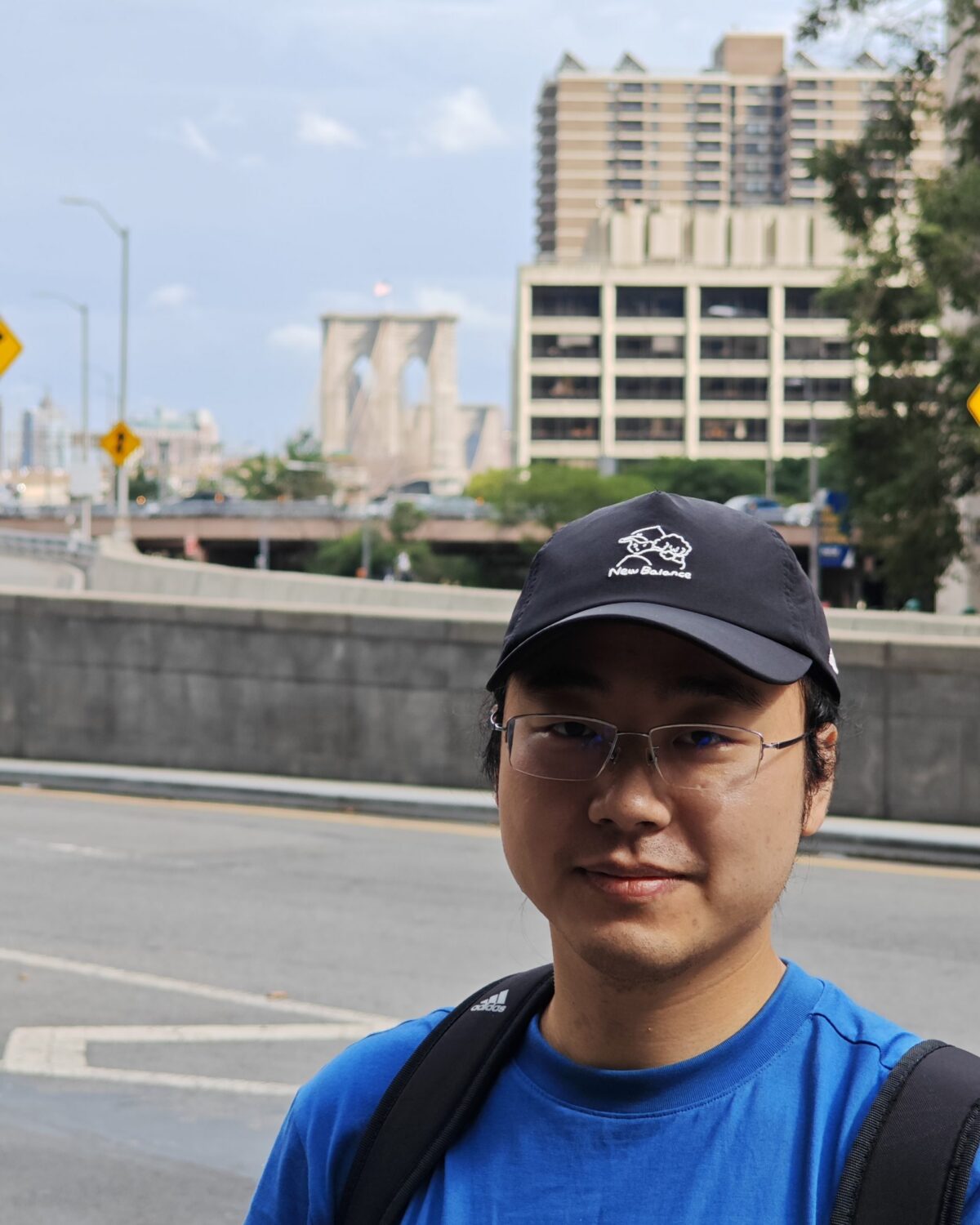 A person wearing glasses, a blue shirt, and a black cap stands outdoors with the Brooklyn Bridge and city buildings visible in the background.