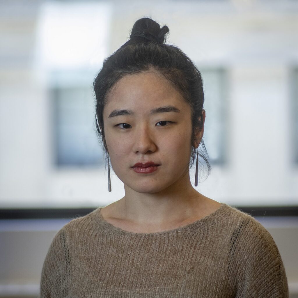A person with long dark hair tied in a bun, wearing a brown sweater and long earrings, stands indoors in front of a blurred window.