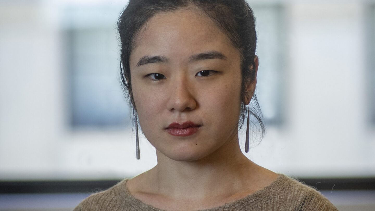 A person with long dark hair tied in a bun, wearing a brown sweater and long earrings, stands indoors in front of a blurred window.