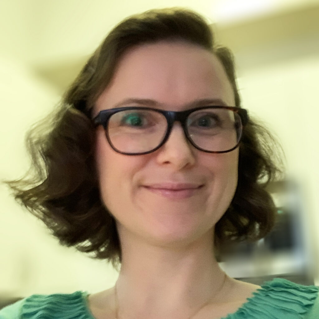 A woman with short wavy brown hair and black glasses smiles at the camera. She is wearing a light green top. The background is softly blurred with warm lighting.