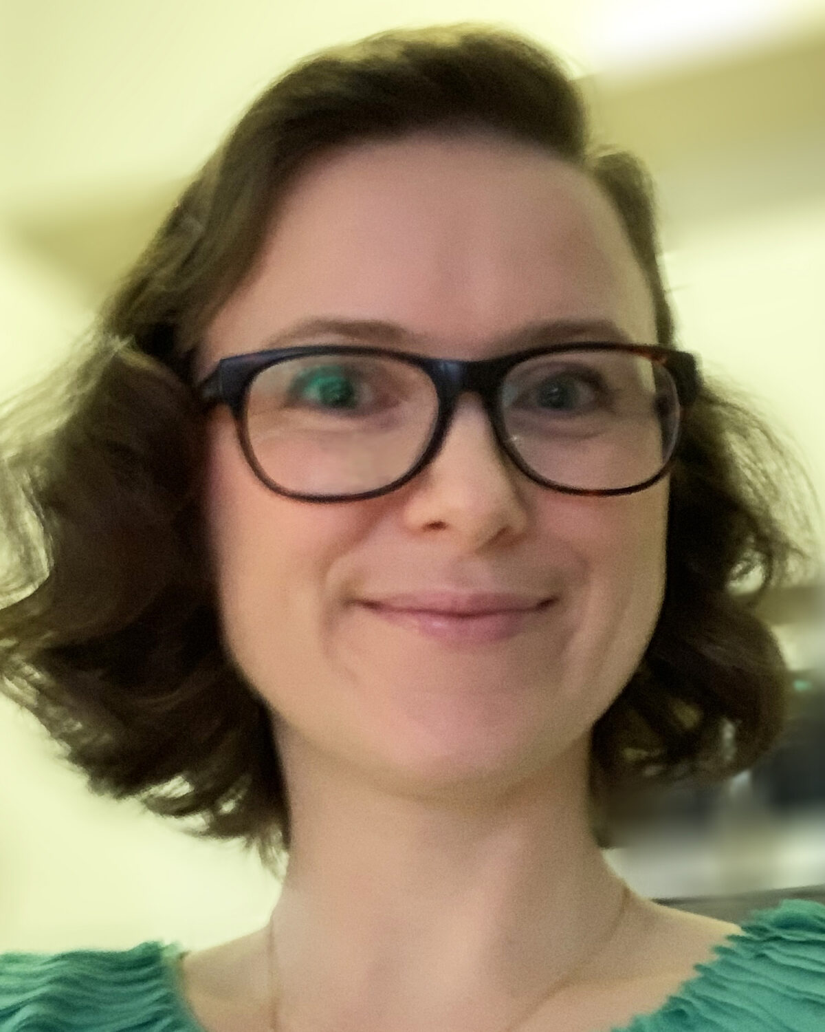 A woman with short wavy brown hair and black glasses smiles at the camera. She is wearing a light green top. The background is softly blurred with warm lighting.