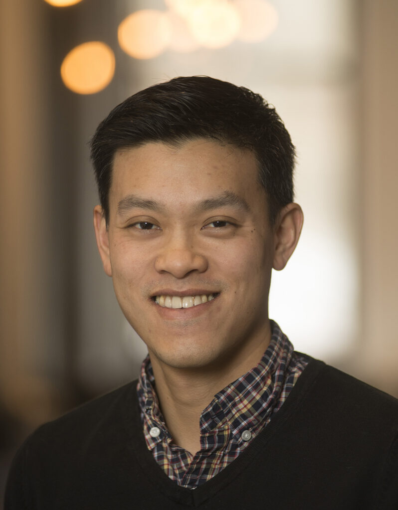 A man with short black hair smiles at the camera. He is wearing a plaid shirt under a dark sweater. The background is softly blurred with warm lights and muted colors.