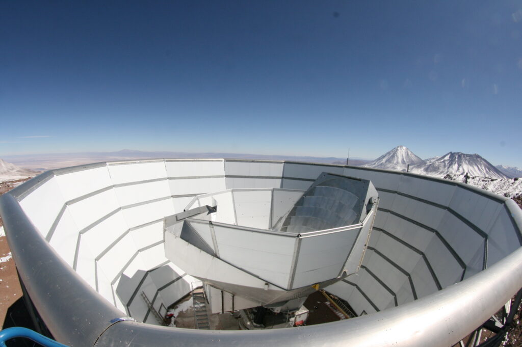 A large white telescope dish sits in a circular structure on a mountain, with a clear blue sky and snow-capped peaks in the background.