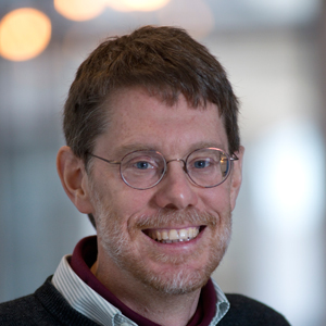 A smiling man with short brown hair, glasses, and a beard is wearing a collared shirt and sweater. The background is softly blurred with warm lights.