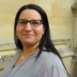 Woman with long dark hair and glasses smiling, wearing a light gray top, standing in front of a tan stone wall.