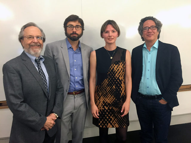 Four people stand together in front of a whiteboard, three men in suits and a woman wearing a patterned dress, all facing the camera and smiling or with neutral expressions.