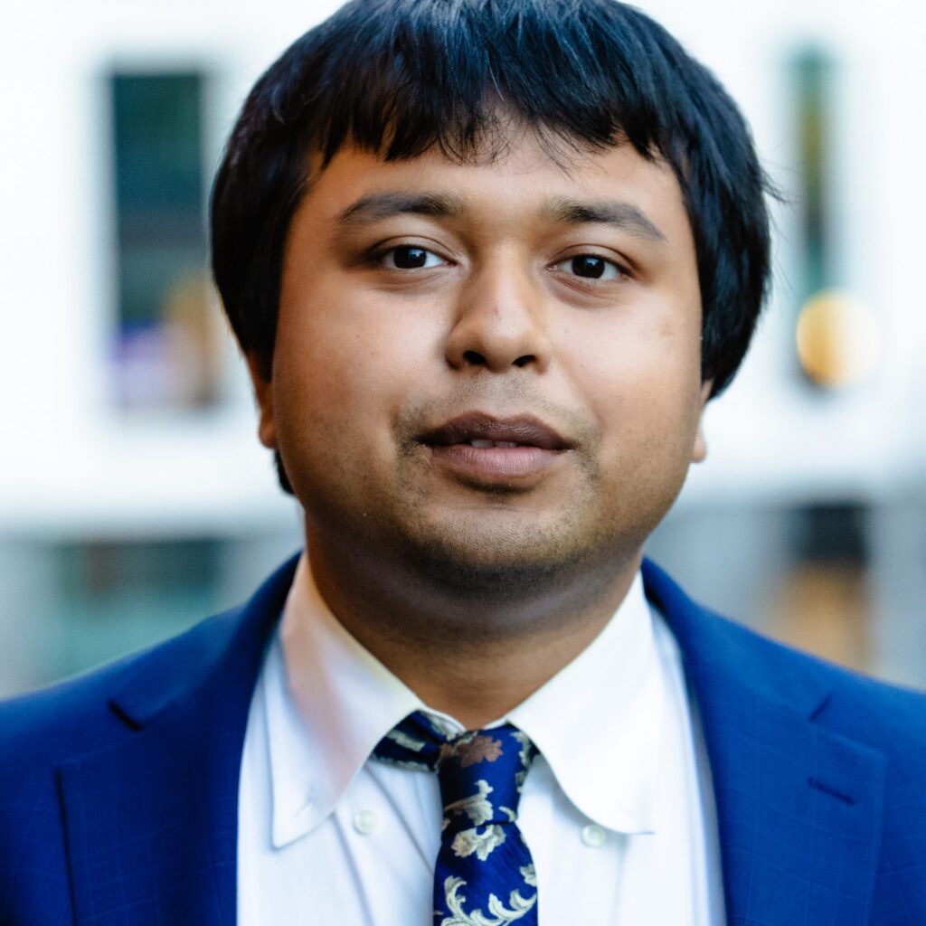 A person with short black hair wearing a blue suit, white shirt, and patterned tie, standing outdoors in front of a blurred building background.