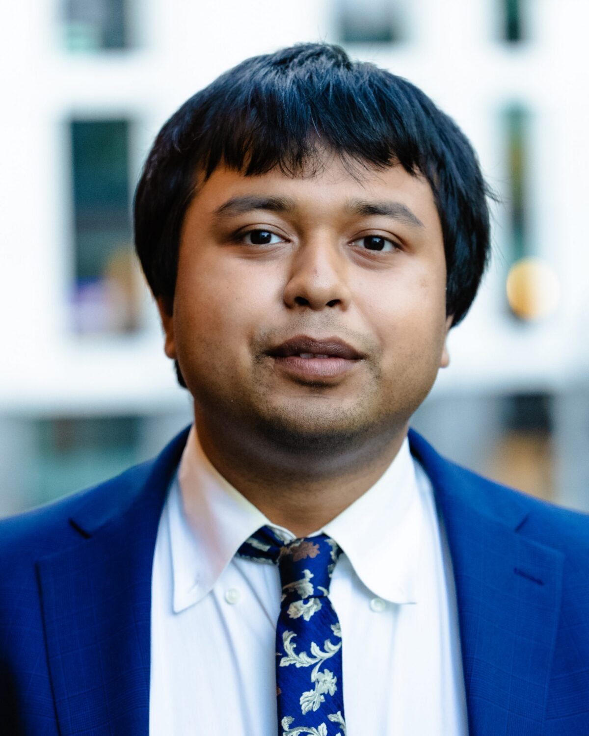 A person with short black hair wearing a blue suit, white shirt, and patterned tie, standing outdoors in front of a blurred building background.
