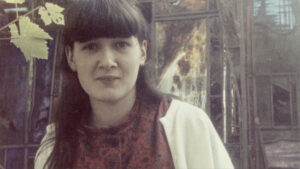 A woman with straight dark hair and bangs stands outdoors, wearing a red patterned top and a light jacket. Green leaves and a glass building with reflections are in the background.