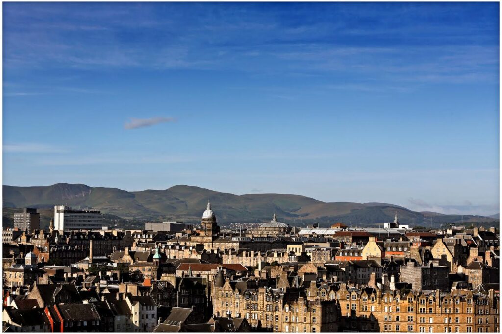 Panoramic view of a historic city with stone buildings, domes, and spires under a clear blue sky, with green hills in the background.