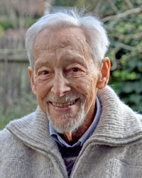 An elderly man with white hair and a beard smiles warmly while wearing a light knit sweater outdoors, with greenery and a wooden fence in the background.