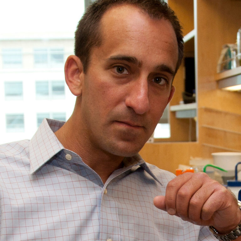 A man in a light plaid shirt sits indoors, looking at the camera with a serious expression, his right hand raised slightly as if emphasizing a point. Shelves and lab equipment are visible in the background.