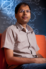 A person with glasses sits on an orange chair in front of a chalkboard covered in mathematical diagrams and equations.