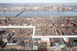 Aerial view of a city with a white path highlighted through rows of brick buildings, trees, and streets, with a river and a distant cityscape in the background.